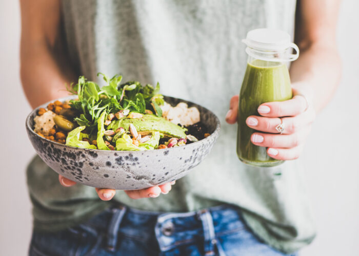 Healthy dinner or lunch. Woman in t-shirt and jeans standing and holding vegan superbowl or Buddha bowl with hummus, vegetable, salad, beans, couscous and avocado and smoothie in hands, square crop